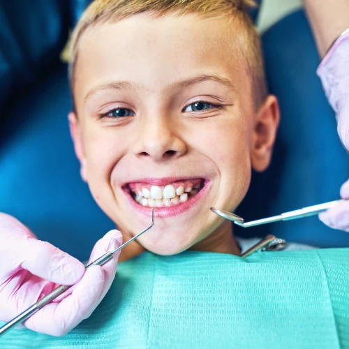 Un bambino biondo sorride mostrando i denti durante un controllo dentistico, mentre il dentista utilizza strumenti odontoiatrici per il trattamento.